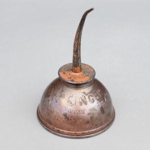 Puede incluir: Lata de aceite Singer vintage con un largo pico. La lata es de metal y tiene un color marrón oxidado. Las palabras "Singer Made in USA" están impresas en el lateral de la lata.