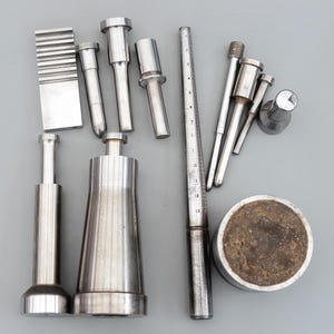 Puede incluir: Un surtido de herramientas de metal plateado, que incluyen varias formas cilíndricas y cónicas, un medidor de anillos y un bloque rectangular texturizado. Las herramientas están dispuestas sobre una superficie gris, mostrando sus diferentes tamaños y diseños.