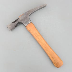 May include: A rock hammer with a steel head and a wooden handle. The hammer head has a flat striking face and a pointed end. The handle is made of light-colored wood.