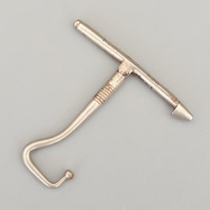 Könnte beinhalten: Ein silberfarbenes Vintage-Werkzeug mit T-förmigem Griff und gebogenem Haken. Der Griff hat eine strukturierte Oberfläche, und das andere Ende hat eine Spitze. Das Werkzeug ist aus Metall und wird wahrscheinlich für einen bestimmten Zweck verwendet.