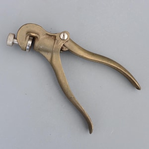 Könnte beinhalten: Ein antikes Säge-Set-Werkzeug aus Messing mit einer silbernen Einstellschraube. Das Werkzeug hat zwei gebogene Griffe und einen kreisförmigen Kopf. Das Säge-Set dient zur Einstellung der Sägezähne. Das Werkzeug liegt auf einer grauen Oberfläche.