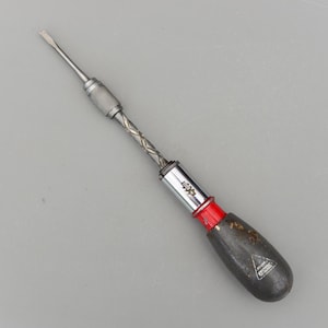 Puede incluir: Un destornillador multi-punta vintage con punta plana. La herramienta presenta un eje de metal plateado con un agarre texturizado, una banda roja y un mango de madera oscura con un logotipo triangular.