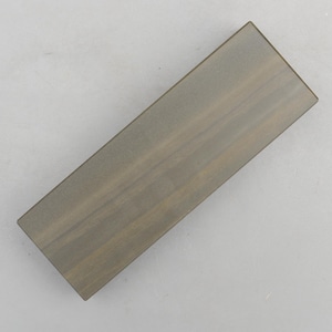 May include: A rectangular sharpening stone with a flat, uniform surface. The stone features a muted, earthy gray-brown color with subtle, darker horizontal grain-like striations running across its length, providing a textured appearance.