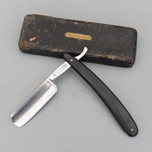 May include: A straight razor with a black handle and a silver blade, alongside a black leather case. The blade is marked with the text "THE FISHER FOR QUALITY".