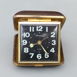 Peut inclure: Un réveil de voyage Timemaster vintage avec un cadran noir, des chiffres blancs et des aiguilles dorées. L'horloge est logée dans un étui en similicuir marron avec un cadre doré. Le cadran indique "Timemaster" et "Alarm".