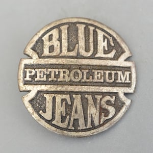 Könnte beinhalten: Ein kreisförmiges, silberfarbenes Metallabzeichen mit den Worten "BLUE PETROLEUM JEANS" in einem Vintage-Schriftzug mit Used-Look geprägt. Das Abzeichen hat ein leicht verwittertes Aussehen.