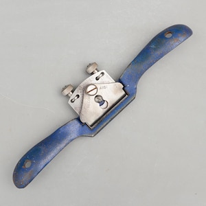 May include: A vintage hand tool, a spokeshave, with a blue painted handle and a silver blade. The tool has two adjustment knobs and a curved design for shaping wood. The metal shows signs of wear, indicating its age and use.