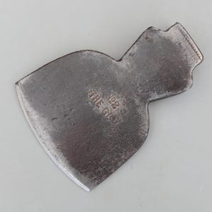 May include: A weathered axe head with a dark, metallic appearance. The axe head features a curved blade and a rectangular top with a textured edge. The text "NO2 THE GEM" is stamped on the surface. This tool is likely used for woodworking or splitting.