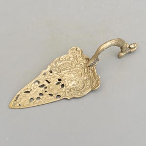 Può includere: Un servizio per torte in metallo dorato con un design ornato e traforato e un manico decorativo. Il manico ha la forma di un uccello con una coda arricciata.