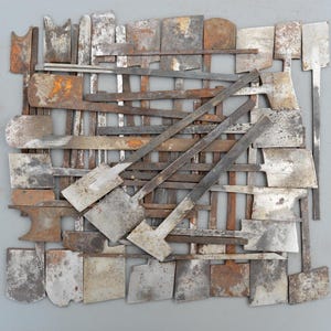 Può includere: Una collezione di vari utensili metallici vintage con lame piatte, rettangolari e curve disposte in una griglia densa e sovrapposta. Gli strumenti in ferro e acciaio mostrano vari gradi di ossidazione, ruggine e texture metalliche grigie.