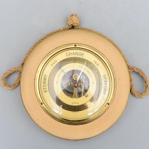 Può includere: Un barometro rotondo vintage con esterno in tessuto beige e quadrante in metallo dorato. Il quadrante presenta una copertura in vetro con un ago che indica le condizioni meteorologiche come "Tempesta" e "Molto secco". Un cordoncino intrecciato è attaccato per appenderlo.