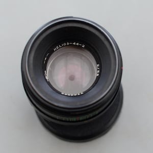 以下が含まれることがあります： シルバーの内部を備えた黒色のHelios-44-2カメラレンズ。レンズの前面には「Helios-44-2」の文字が印刷されています。レンズの側面には、緑、赤、白のマークが並んでいます。