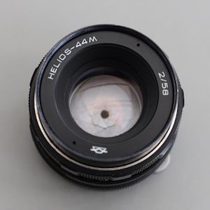May include: A black Helios-44M 2/58 camera lens with silver detailing. The lens features a circular design with the text "HELIOS-44M" and "2/58" printed on the outer ring. The lens is designed for photography.