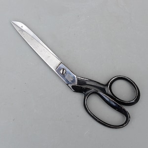 May include: A pair of silver and black scissors. The blades are silver and the handles are black. The scissors are lying on a gray surface. The scissors are likely used for cutting fabric or paper.