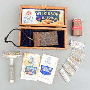 Może przedstawiać: Zestaw do golenia Wilkinson Sword z epoki w drewnianym pudełku z niebieskim aksamitnym wykończeniem. Zestaw zawiera srebrną maszynkę do golenia, skórzane etui, pięć ostrzy i dwie instrukcje obsługi. Pudełko jest oznaczone "Wilkinson Razor Empire Model Lift 10/6".