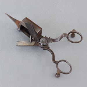 May include: An antique metal tool, resembling scissors, with a complex design. It has a rectangular body, a pointed blade, and two circular finger holes. The metal has a dark, aged appearance, suggesting its vintage.