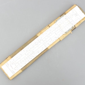 May include: A vintage slide rule in a cardboard case. The slide rule is white with black markings and a metal slider. The case is tan and rectangular, with a partially visible label.