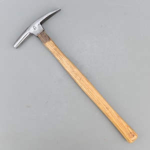 May include: A metal and wood geological hammer with a pointed end and a flat striking surface. The hammer has a light brown wooden handle and a silver-colored metal head. The tool is designed for breaking rocks and geological work.