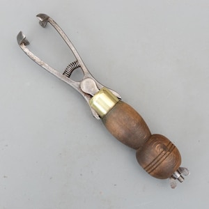 May include: A vintage metal and wood tool with a spring and a wooden handle. The tool has a brass collar and a screw at the end of the handle. The tool is likely used for leatherworking or a similar craft.