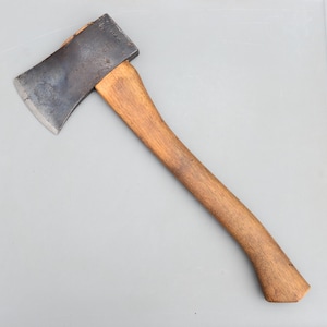 May include: A hand axe featuring a dark, weathered metal head with a visible stamped logo and a curved, light brown wooden handle. The metal blade shows signs of use with a worn finish, while the wooden handle has a smooth, natural grain texture.