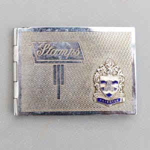 May include: Silver-coloured metal stamp case with a textured surface. The case features the word "Stamps" in a stylized font and a crest with the word "Paignton" in blue. The case is rectangular and hinged.