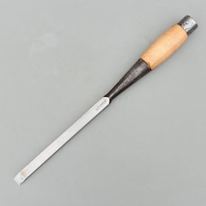 May include: A steel wood chisel featuring a long, flat metal blade with a beveled edge and a light-colored wooden handle. The metal neck connects the blade to the handle, which is capped with a dark metal ferrule. The blade is stamped with the text "F. HERDER A. SN" and "SHEFFIELD".