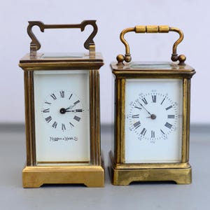 Könnte beinhalten: Zwei antike Messing-Reiseuhren mit weißen Zifferblättern und römischen Ziffern. Die Uhren haben eine rechteckige Form mit einem Griff oben. Eine Uhr trägt den Text "Mappin & Webb" auf dem Zifferblatt.