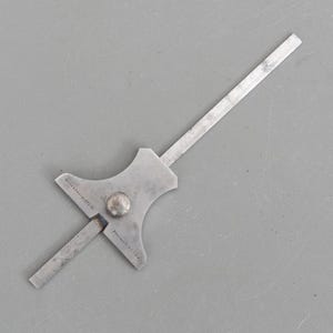 Peut inclure: Jauge de profondeur en métal vintage, avec une lame plate et allongée et un corps plus large et stylisé. Le corps comporte un rivet circulaire et le texte "Brown & Sharpe Mfg. Co." et "Providence, R.I. U.S.A.". L'outil est argenté et semble bien utilisé.