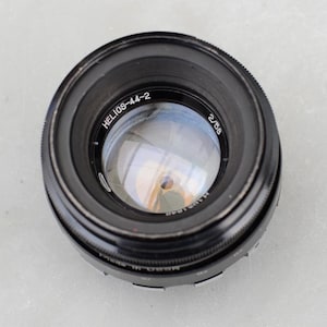 May include: A black Helios-44-2 camera lens. The lens has the text "Helios-44-2" and "2/58" printed on it. The lens has a reflective glass surface and a ridged focusing ring.