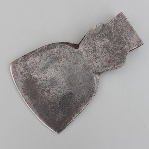 Puede incluir: Una cabeza de hacha antigua y desgastada con una hoja ligeramente curvada. El metal tiene un aspecto oscuro y envejecido con desgaste visible, lo que sugiere su uso a lo largo del tiempo. La cabeza del hacha es ancha y plana, con un filo distintivo.