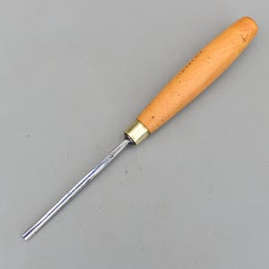 Può includere: Uno scalpello per intaglio del legno con un manico in legno marrone chiaro, una ghiera in ottone e una lama color argento. Lo scalpello è progettato per lavori di falegnameria e intaglio dettagliati.