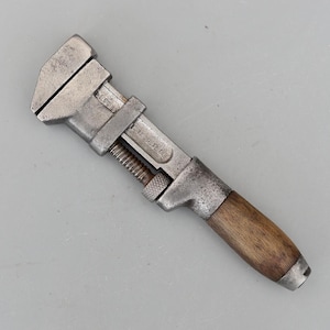 May include: A vintage adjustable wrench with a metal head and a wooden handle. The wrench is silver-toned with visible wear and tear, and the handle is a warm brown colour. The tool is marked "U.S.A."