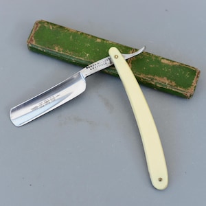 Puede incluir: Una navaja de afeitar recta vintage con mango color crema y hoja plateada. La hoja está marcada con la palabra "EMU" y "FORDA & HALLS SHEFFIELD". La navaja está abierta y apoyada sobre un estuche rectangular verde.