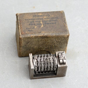 May include: A vintage metal numbering machine with a serial number, in its original box. The machine has a series of numbered wheels and a lever to advance the numbers. The box is made of cardboard and has the brand name "PERMOID" printed on it.