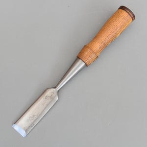 May include: A woodworking chisel featuring a light brown wooden handle and a silver-coloured blade. The handle has a darker brown end. The blade is rectangular with a slightly curved edge. This chisel is designed for detailed woodworking.