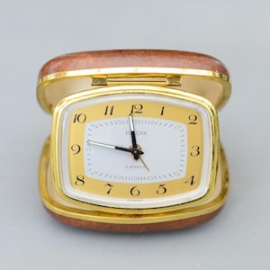 May include: A vintage Europa travel alarm clock in a brown leather case. The clock face is gold with black numerals and hands. The text "EUROPA" and "2 JEWELS" are visible. The clock is open, showing the time.