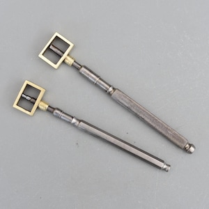 May include: Two antique metal tools with brass square heads and textured handles. The tools have a screw mechanism and a dark, metallic finish. The handles are cylindrical and hexagonal. The tools are likely used for a specific craft or repair.