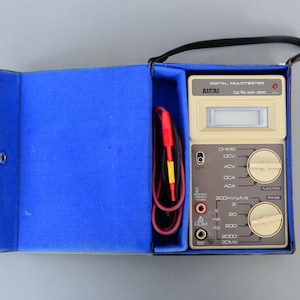 May include: A beige digital multimeter with a black carrying case. The multimeter has a digital display, a function knob, and a range knob. The text on the multimeter reads "DIGITAL MULTITESTER", "AITAI", "Cat No. KD-300", "OHMS", "DCV", "ACV", "DCA", "ACA", "200mV/A/I", "FUNCTION", "RANGE", "2", "20", "A", "mA", "COM", "200", "2000", "20MII".