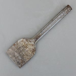 May include: A vintage, weathered metal chisel with a wide, rectangular blade and a long, rectangular handle. The handle is stamped with the text "SPEAR AND JACKSON ENGLAND". The tool has a worn, aged appearance.