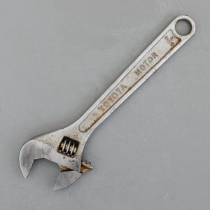 May include: A silver adjustable wrench with the text "TOYOTA MOTOR" printed on the handle. The wrench has a rust-colored patina and a circular hole at the end of the handle. The jaws are open, ready for use.