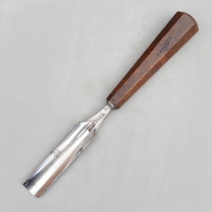 May include: A woodworking chisel with a polished steel blade and a dark brown wooden handle. The handle is octagonal in shape. The blade has text etched into the metal. The chisel is designed for shaping and carving wood.