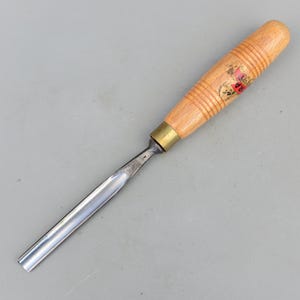 Puede incluir: Un formón de talla de madera con un mango de madera marrón claro y una hoja de metal. El mango tiene una virola de latón y un logotipo rojo y dorado. La hoja es curva y tiene una superficie pulida y reflectante.