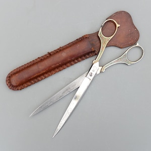 以下が含まれることがあります： 銀色の刃と装飾的なハンドルが付いた開いたハサミと、茶色の革製の鞘。ハサミには金と赤の装飾が施されています。鞘は、ステッチされたエッジを持つ濃い茶色です。ハサミの長さは約20cmです。