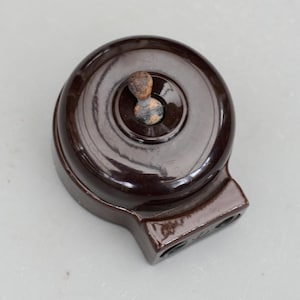 May include: A vintage, dark brown light switch with a circular design and a toggle switch. The switch has a rectangular base with two holes. The toggle switch is made of a lighter brown material.