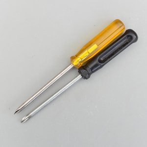 Może przedstawiać: Dwa śrubokręty ze srebrnymi trzonkami i różnokolorowymi uchwytami. Jeden ma przezroczysty żółty uchwyt, a drugi czarny. Końcówki śrubokrętów są przeznaczone do śrub krzyżakowych.