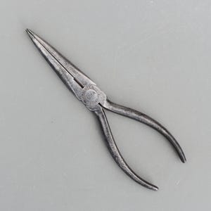 Op de afbeelding: Een paar lange punttangen met een donkere, metalen afwerking. De tang heeft een slanke, puntige punt en gebogen handgrepen. De tekst "KRAEUTER USA" is op het scharnier gestempeld. De tool is ontworpen voor precisiewerk.