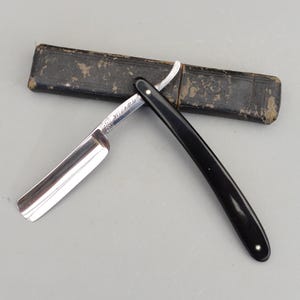 以下が含まれることがあります： 光沢のある銀色の刃と黒い柄のヴィンテージストレートカミソリ。 カミソリは開いており、鋭い刃先が見え、使い古された濃い革のケースの隣に置かれています。 「WIZARD」の文字が刃に刻まれています。