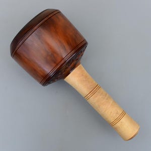 May include: A wooden mallet with a dark brown head and a light brown handle. The head has decorative carved details and the handle has three carved lines. The mallet is angled against a gray background.