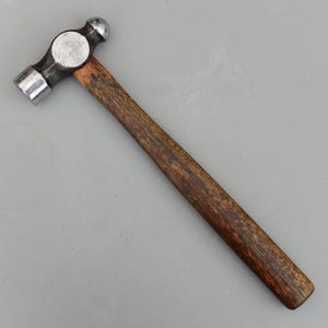 May include: A ball-peen hammer with a dark wooden handle and a metal head. The hammer head has a flat face on one side and a rounded, ball-shaped face on the other. The handle is a dark brown color.