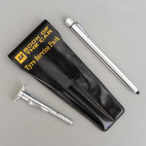 Peut inclure: Un étui en cuir noir "Tyre Service Pack" avec le texte "AA BOOK OF THE CAR" en jaune. Un manomètre de pression des pneus et un stylo argenté sont également visibles.